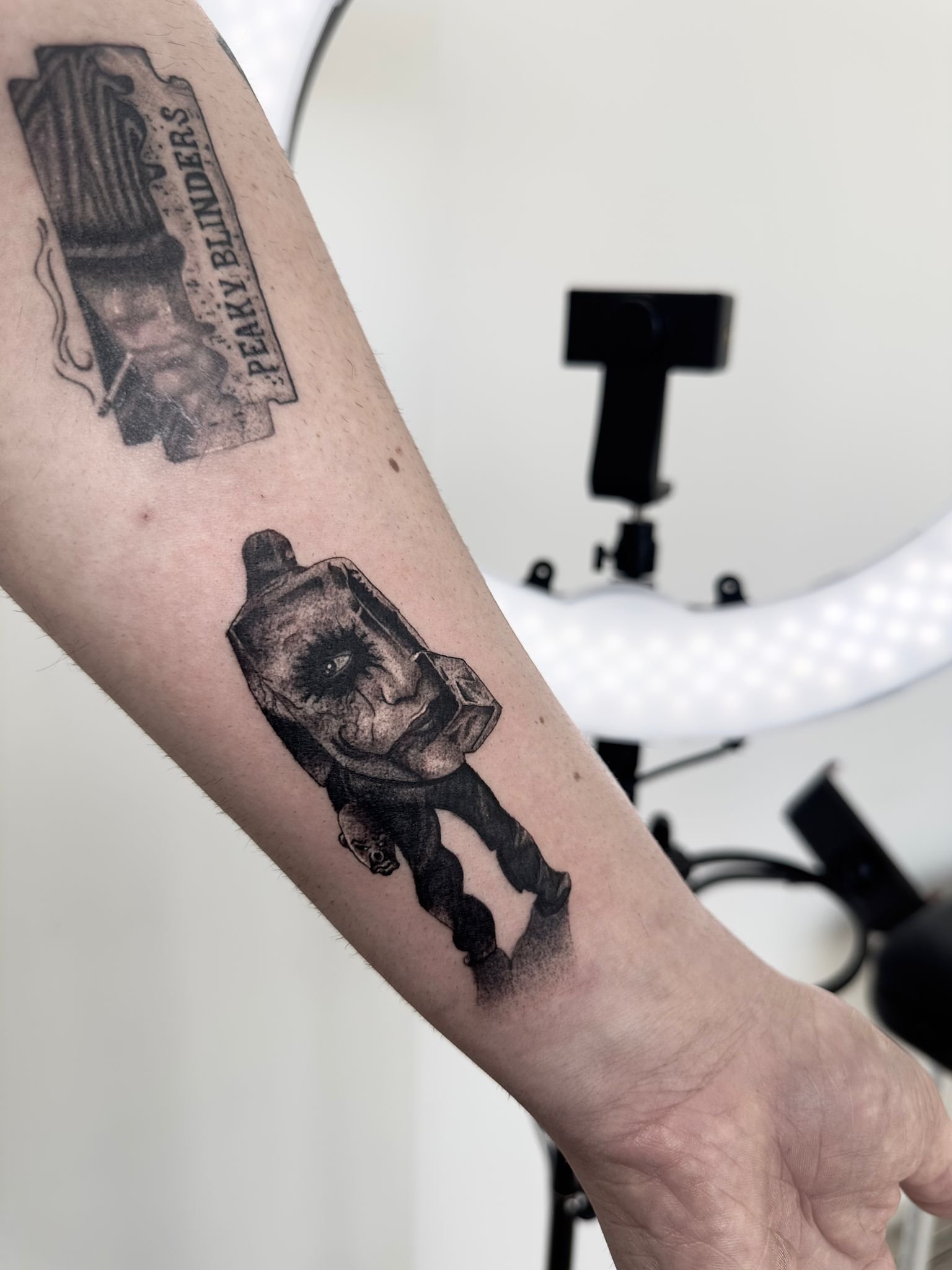 Tatuaje realista en negro y gris de figura enmascarada en el antebrazo, con sombreado y detalles finos.