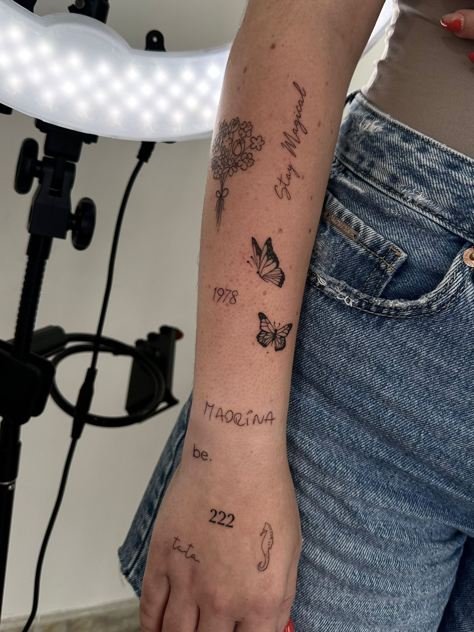 Conjunto de tatuajes fineline con lettering y símbolos (ramo, “Stay Magical”, mariposas, 1978, 222, “MADRINA”, “be.”, “tata”, caballito de mar) en antebrazo y mano.